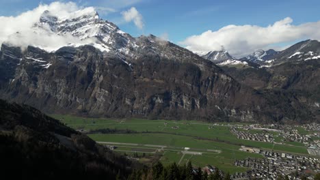 Aerial-drone-shot-flying-over-a-small-town-along-the-valley-surrounded-by-snow-covered-mountain-peaks-in-Switzerland-at-daytime