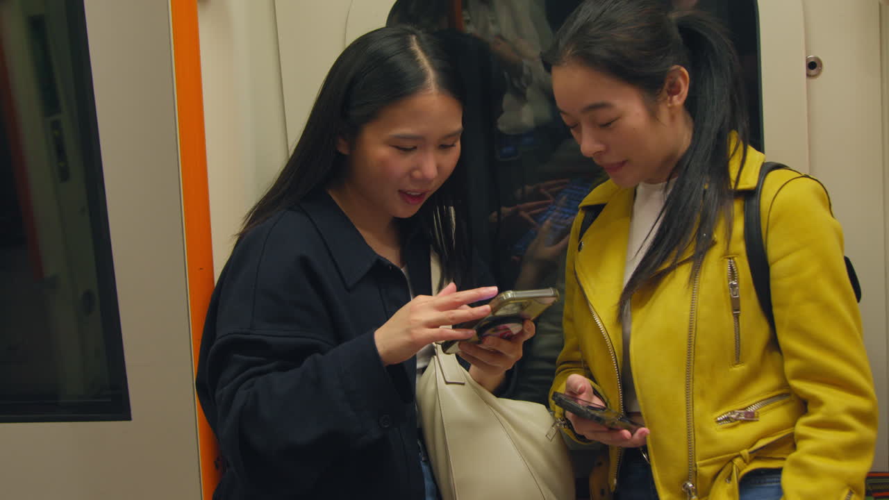 Two Young Female Friends Standing In Carriage On Underground Train Looking At Mobile Phones 1