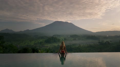 blond caucasian woman in bikini sitting on infinity pool edge overlooking magical mount agung at dawn