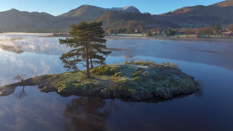 Kleine-Insel-In-Einem-Ruhigen-See-Mit-Spiegelungen-Und-Einer-Umlaufbahn,-Die-Nebel-Auf-Der-Wasseroberfläche-Und-Hellen-Sonnenaufgang-Erkennen-Lässt