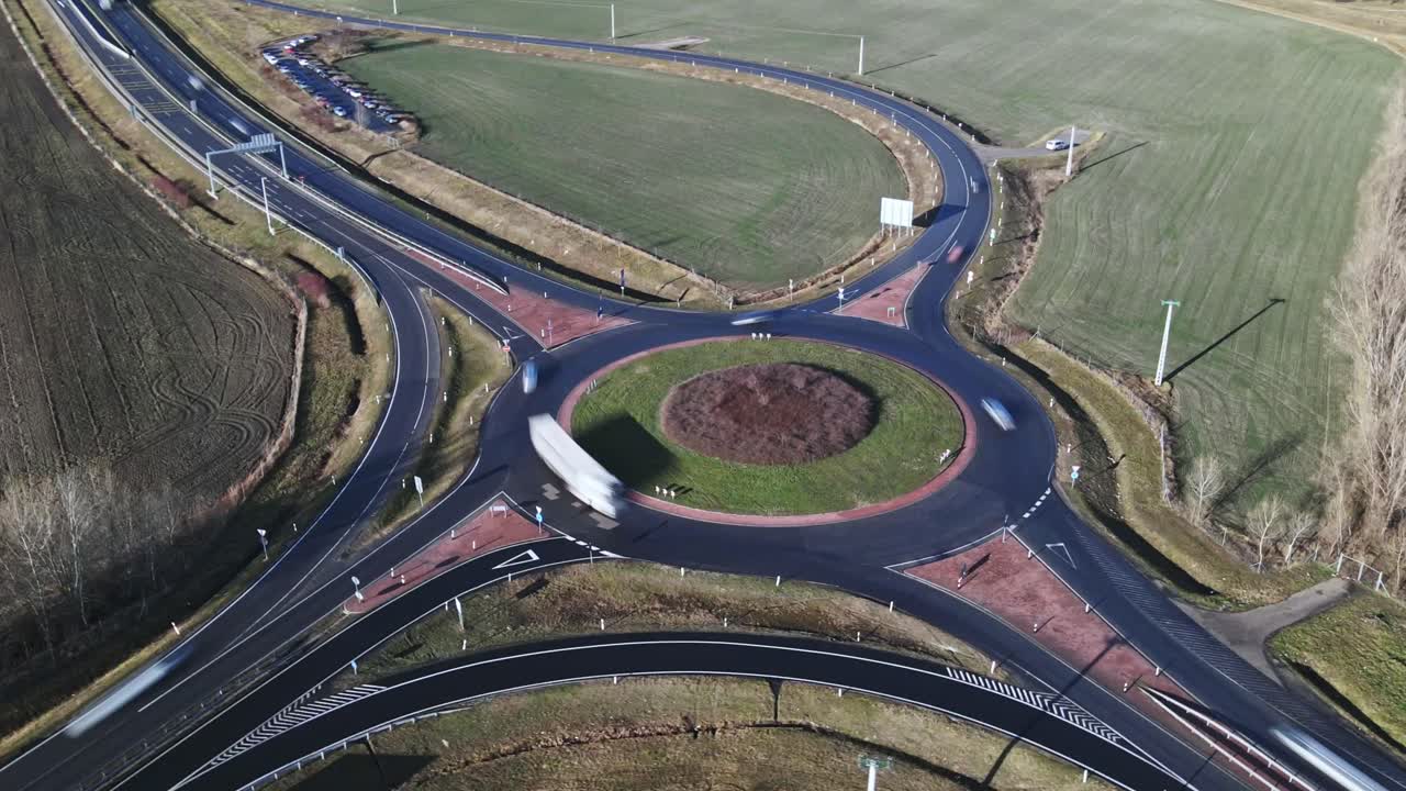 An aerial time-lapse video of a roundabout with cars and trucks moving at high speed. The video captures a well-structured road network, connected highways in the background.