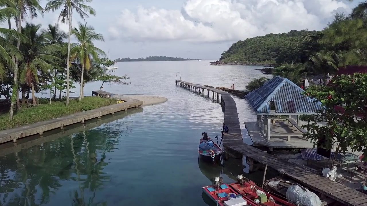 Trên một hòn đảo nhiệt đới, drone bay qua một con suối hướng ra biển, bạn có thể thấy những chiếc thuyền nhỏ neo đậu gần cầu tàu. Cây cọ ở một bên, một bãi cát nhỏ nơi con suối đổ ra biển.