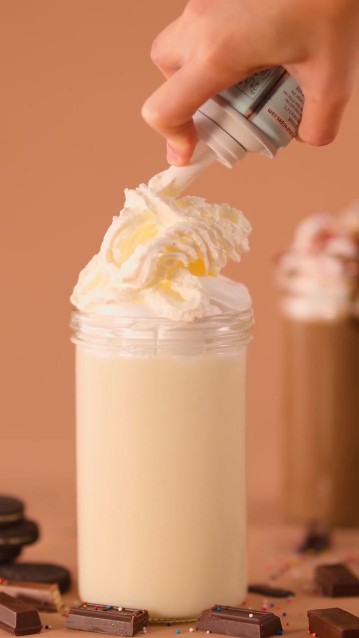 Primo piano di una mano che guarnisce un milkshake alla vaniglia con panna montata, biscotti, cioccolato, illuminazione calda da studio.