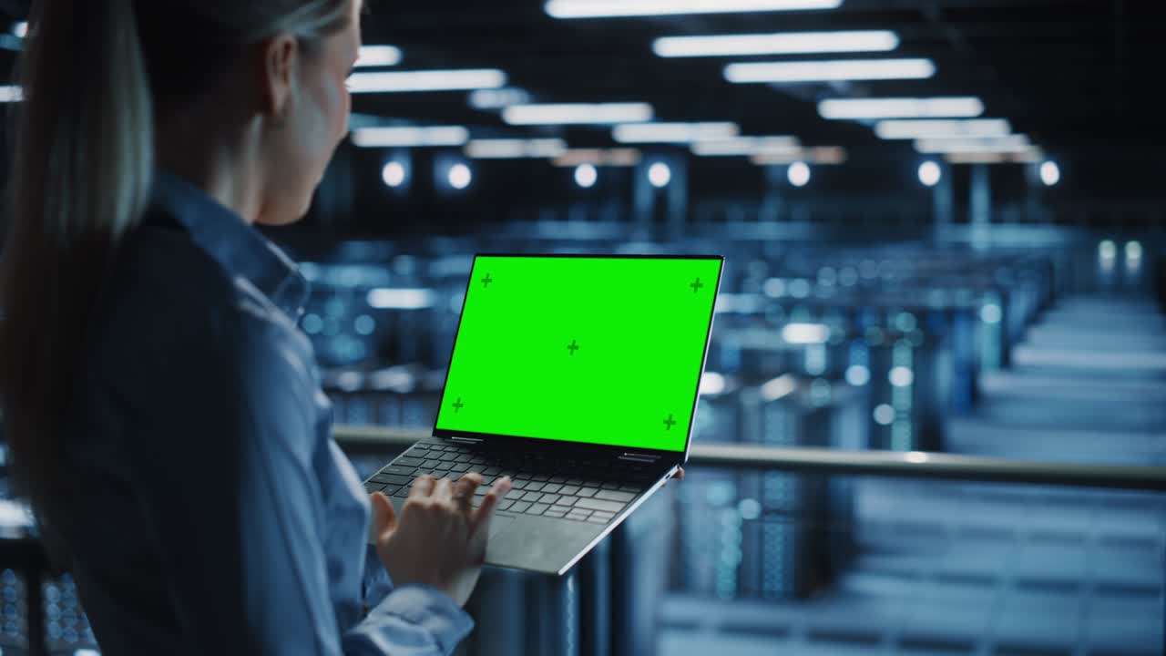Female It Specialist Using Chroma Key Laptop in Data Center. Big Server Farm Cloud Computing Facility with Maintenance Engineer Working with Green Screen Computer. Medium over the shoulder Shot