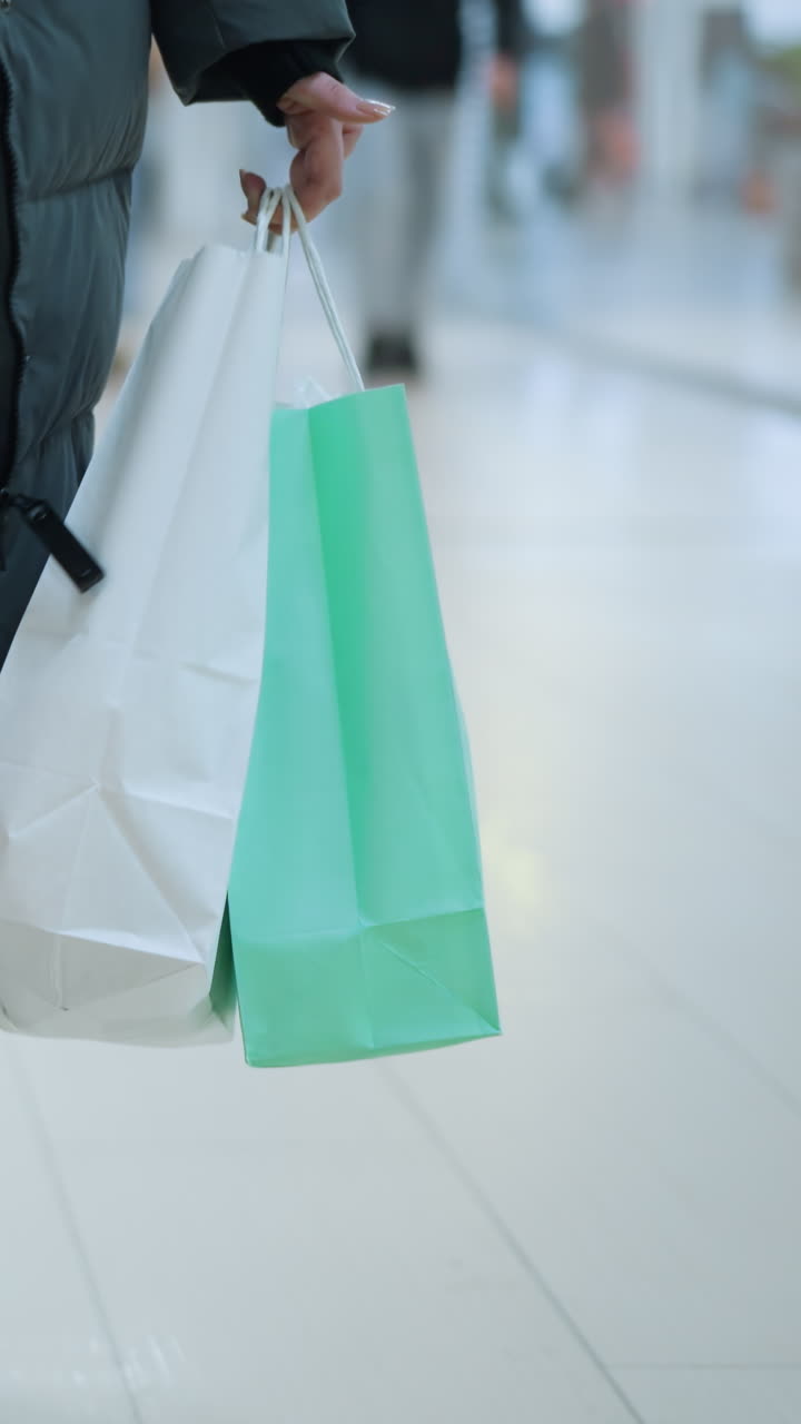 dama en pantalones vaqueros y chaqueta negra caminando en un espacio comercial bien iluminado llevando un bolso negro y dos bolsas de compras, una blanca y una verde menta, fondo borroso con otros compradores