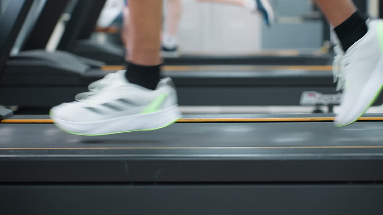 lower angle leg view of two people running on treadmills in bright gym, white sneakers in motion, blurred seated figures and equipment in background, dynamic workout scene