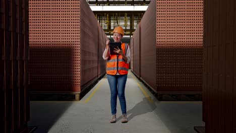 full body of asian female engineer with safety helmet taking note on the tablet and looking around while standing with red brick packed in stacks are stored