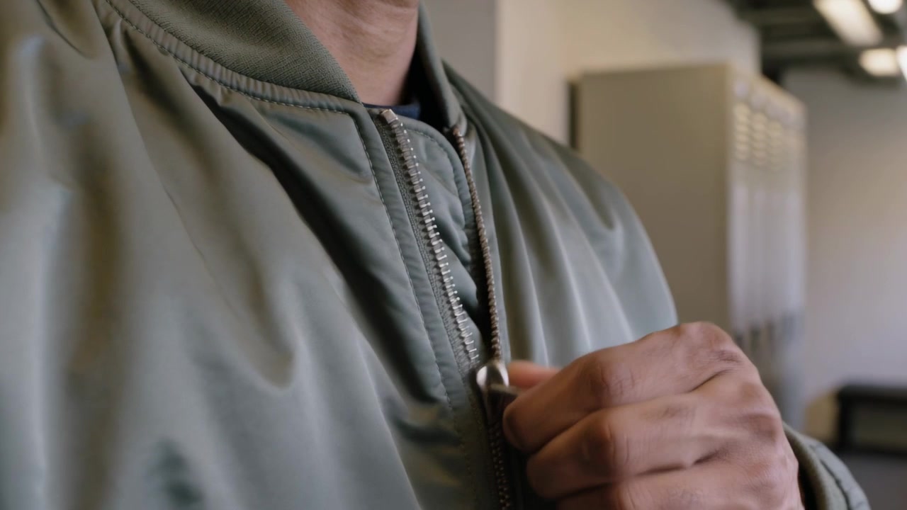 Reaching right hand, man pulling zipper up in locker room, closing olive-green bomber jacket