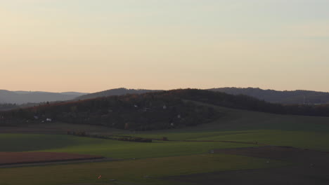 Eine-Zeitrafferansicht-Eines-Natürlichen-Flughafenfeldes-Und-Eines-Hügels,-Wo-Man-Mehrere-Kleinere-Häuser-Sehen-Kann,-Die-Unter-Einem-Grat-Gebaut-Wurden,-Aufgenommen-Während-Des-Sonnigen-Herbstsonnenuntergangsgebiets-Von-Brünn-In-Der-Tschechischen-Republik