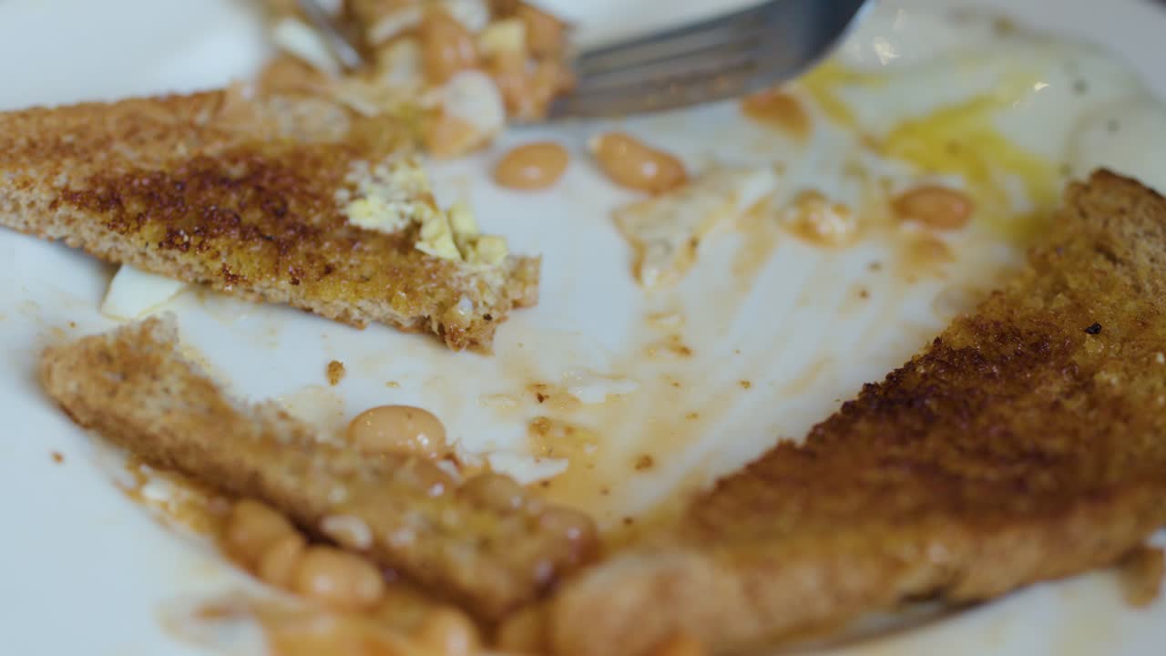 Close-up of fork and knife cutting toast, eggs, and beans on white plate, natural lighting