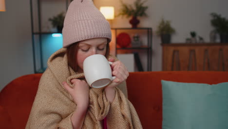 Sick-children-girl-wear-hat-wrapped-in-plaid-sit-alone-shivering-from-cold-on-sofa-drinking-hot-tea