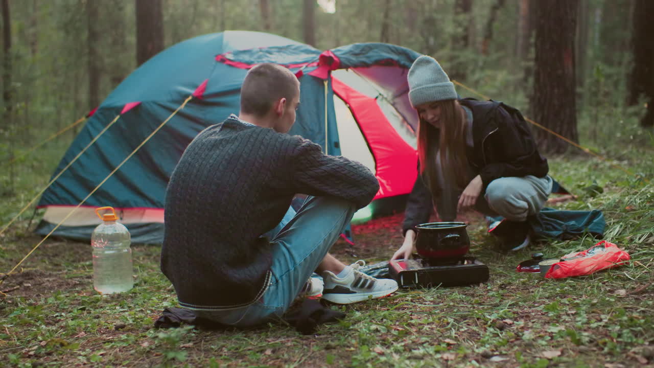 young tourists camping in forest with woman cooking on portable stove while man watches beside tent during outdoor adventure surrounded by trees gear and forest floor