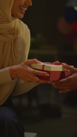 Vertical-Video-Of-Woman-Giving-Female-Friend-Birthday-Present-At-Home-With-Party-Balloons-In-Background-1