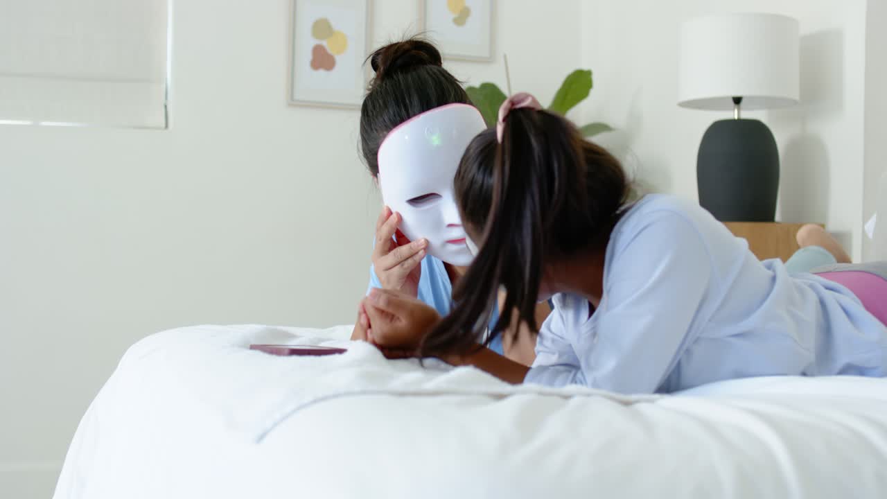Mamma e figlia di diverse etnie sul letto, la mamma solleva una maschera LED, la figlia la tocca, testando la maschera con il telefono