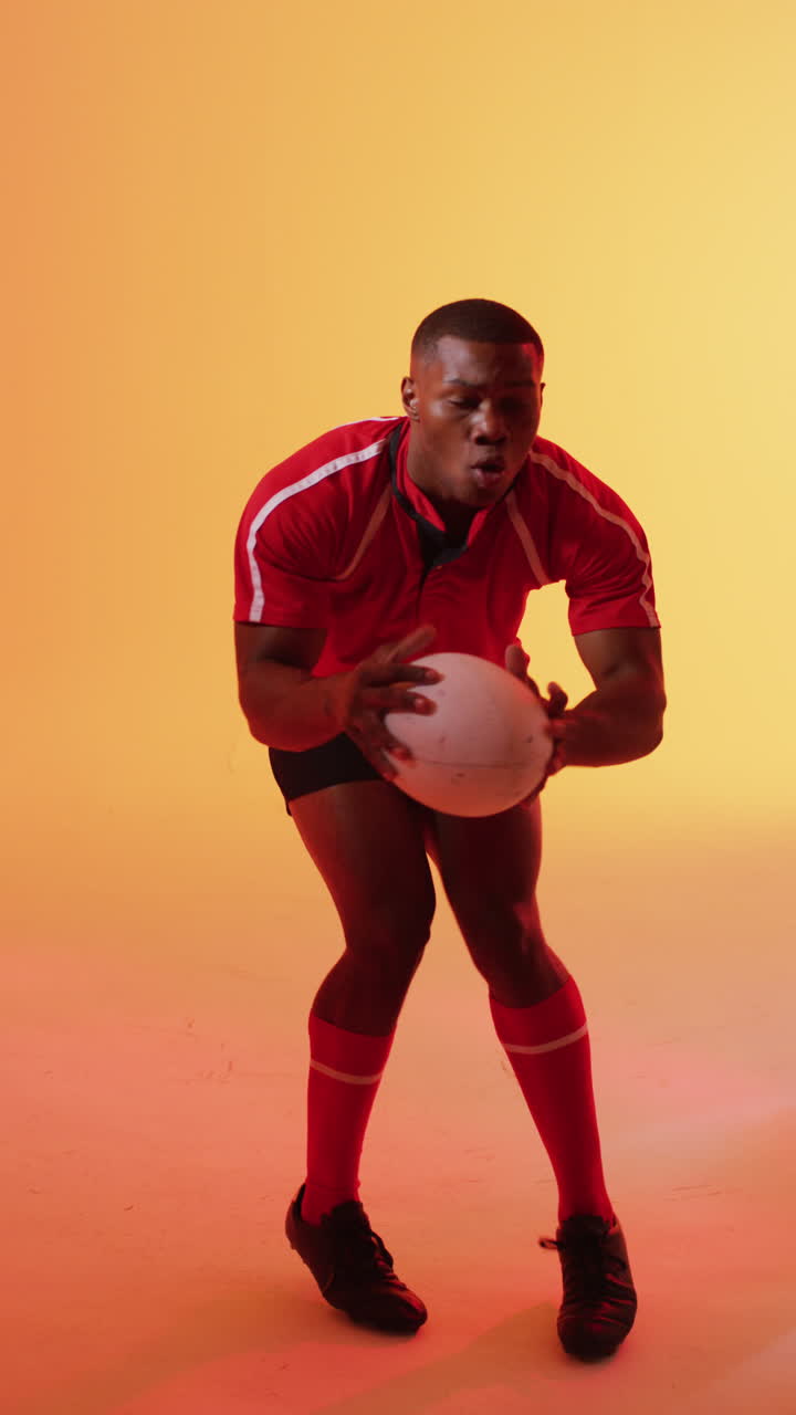 Vertical video of african american male rugby player catching rugby ball over pink lighting