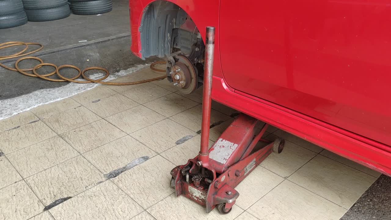 A small red car is seen in a workshop, undergoing a tire change. The front tire is replaced as the car is slightly lifted for ease.