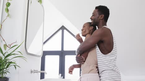 Happy-african-american-couple-brushing-teeth-and-embracing-in-bathroom,-slow-motion