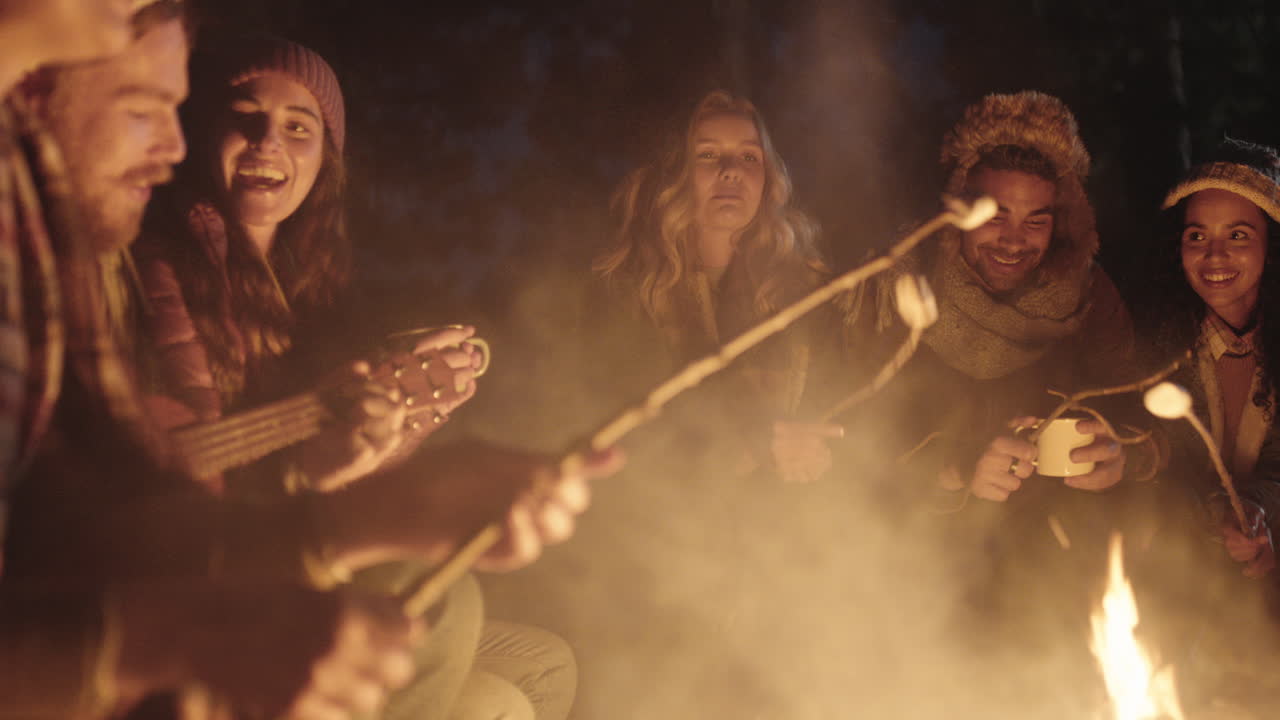 amigos felices acampando sentados junto a una fogata en el bosque por la noche charlando bebiendo chocolate caliente asando malvaviscos compartiendo calidez disfrutando de una aventura al aire libre