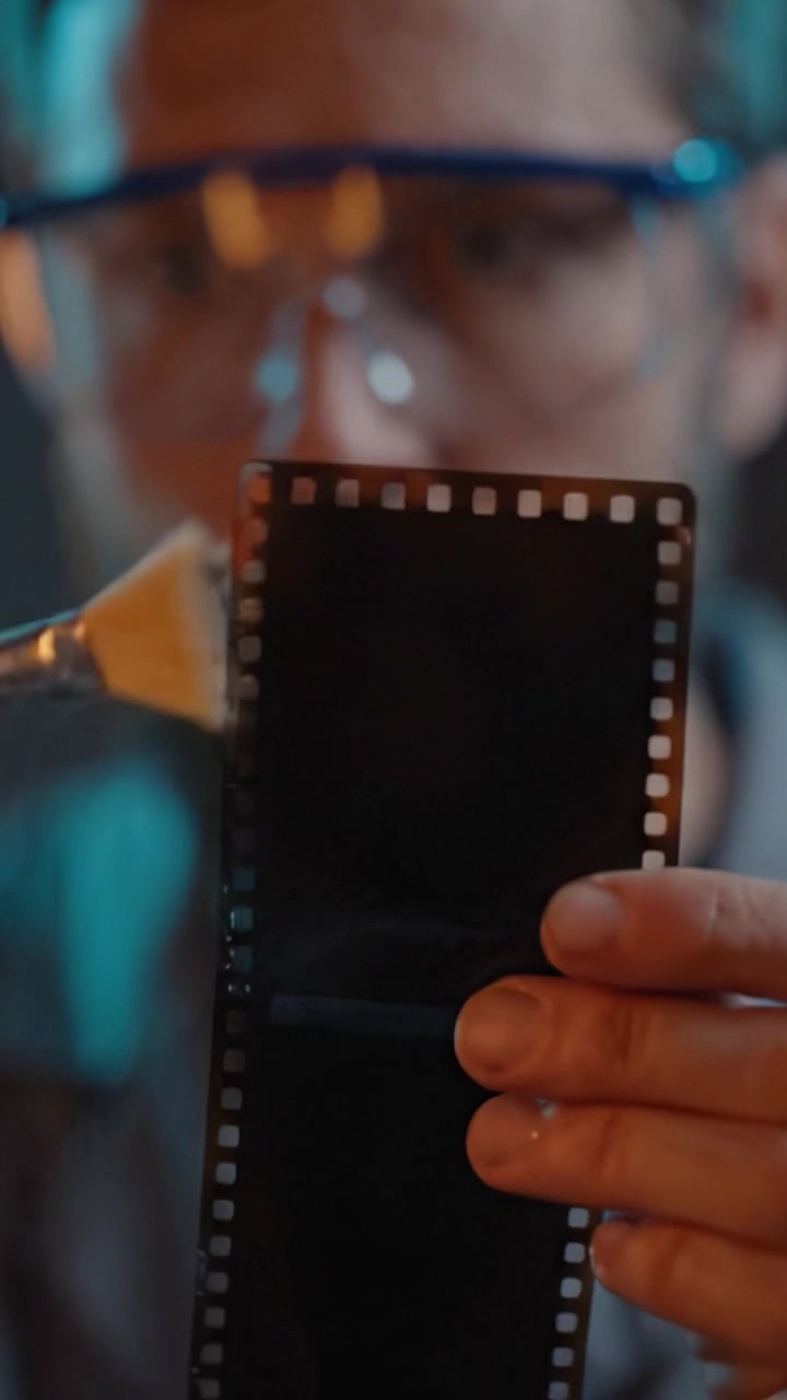 Bir fırça kullanarak bir fotoğraf film şeridini titizlikle temizleyen, film koruma ve bakım tekniklerinin ince detaylarını sergileyen odaklanmış bir kişi.