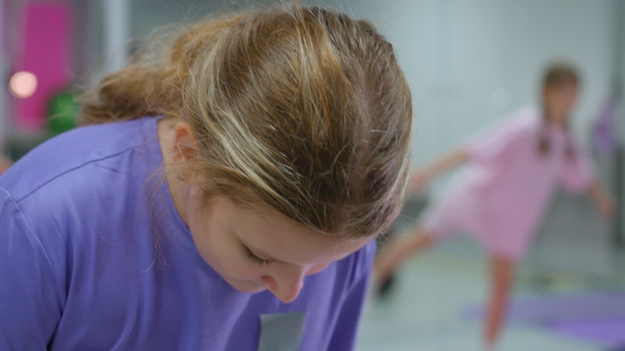 Close-up of young girl in purple outfit concentrating on something in front of her while a blurred child in pink in the background attempts to balance on one leg during a fitness training session