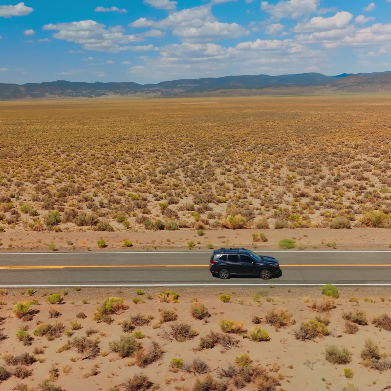 Cars going by the empty road in the sunny desert. Mountainous horizon line under blue cloudy sky at backdrop