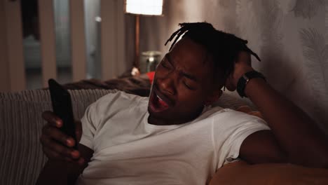 Young-black-male-relaxing-on-the-sofa-using-a-smart-phone-and-yawning-out-of-tiredness