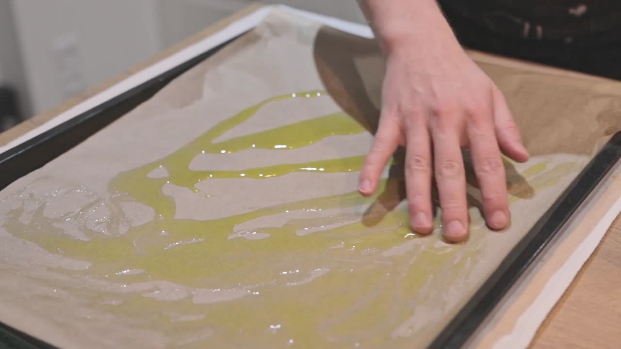 Primo piano di mani che versano olio d'oliva su carta da forno adagiata su una teglia in metallo. Preparazione della teglia per l'impasto della focaccia. Inserto tutorial di cucina incentrato sulla texture