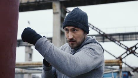 close-up view of caucasian man in black beanie hitting a punching bag outdoors an abandoned factory on a cloudy morning