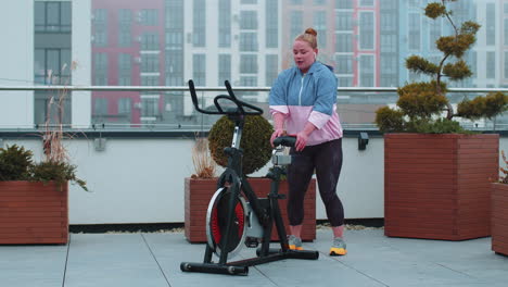 Healthy-Caucasian-woman-exercising-workout-on-stationary-cycling-machine-bike-on-house-rooftop-gym