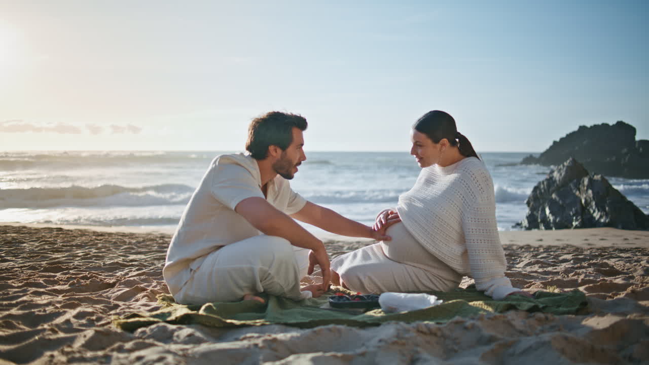 저녁 피크닉 임신 한 커플이 해변 모래에 앉아 로맨틱한 데이트를 보내고 있습니다.