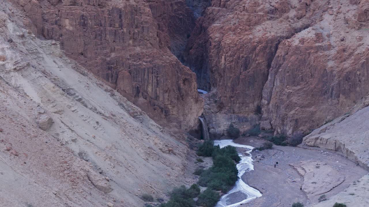 Ripresa aerea con drone di una cascata di acqua torrenziale fangosa che si riversa nel profondo canyon del fiume Kidron nel deserto della Giudea, Israele, con minuscole figure di escursionisti che osservano il raro fenomeno invernale