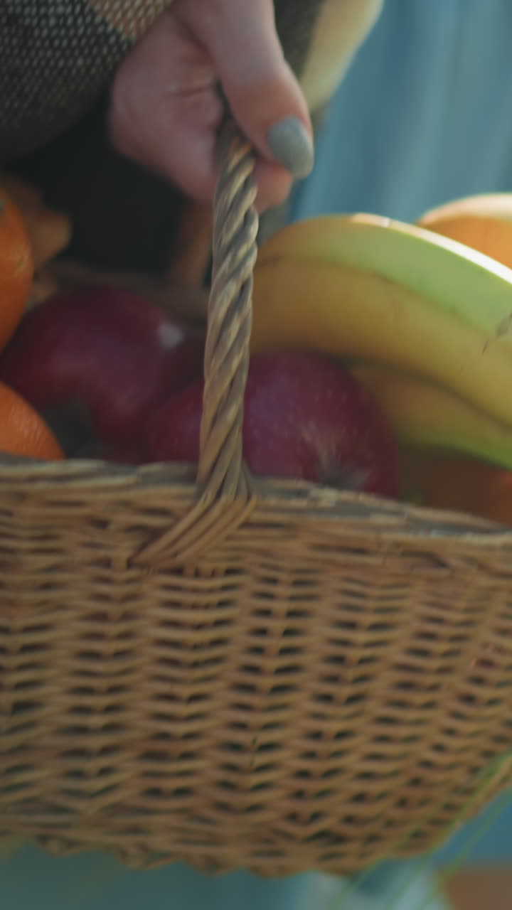 A woman in a blue gown carries a basket of fresh fruit, including bananas, oranges, and apples, in a sunlit field, with a child faintly visible in the background
