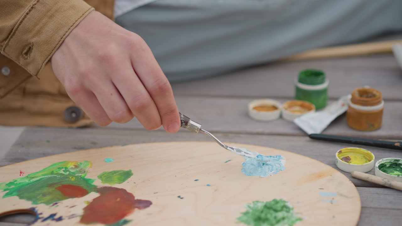 close up hand mixing vivid paints on round palette, muralist energy captured in quick palette knife motion, jars and tubes of pigment spill on table, bold color testing and expressive rhythm, studio