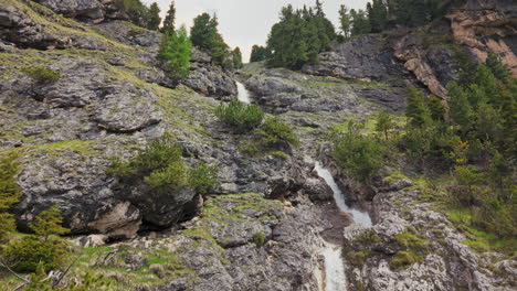 Close-up-aerial-view-of-twin-waterfalls-cascading-over-rocky-terrain-in-the-Dolomite-mountains,-surrounded-by-greenery