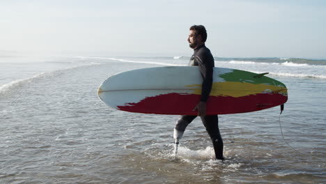 side view of a male surfer with bionic leg coming out of ocean holding surfboard under arm