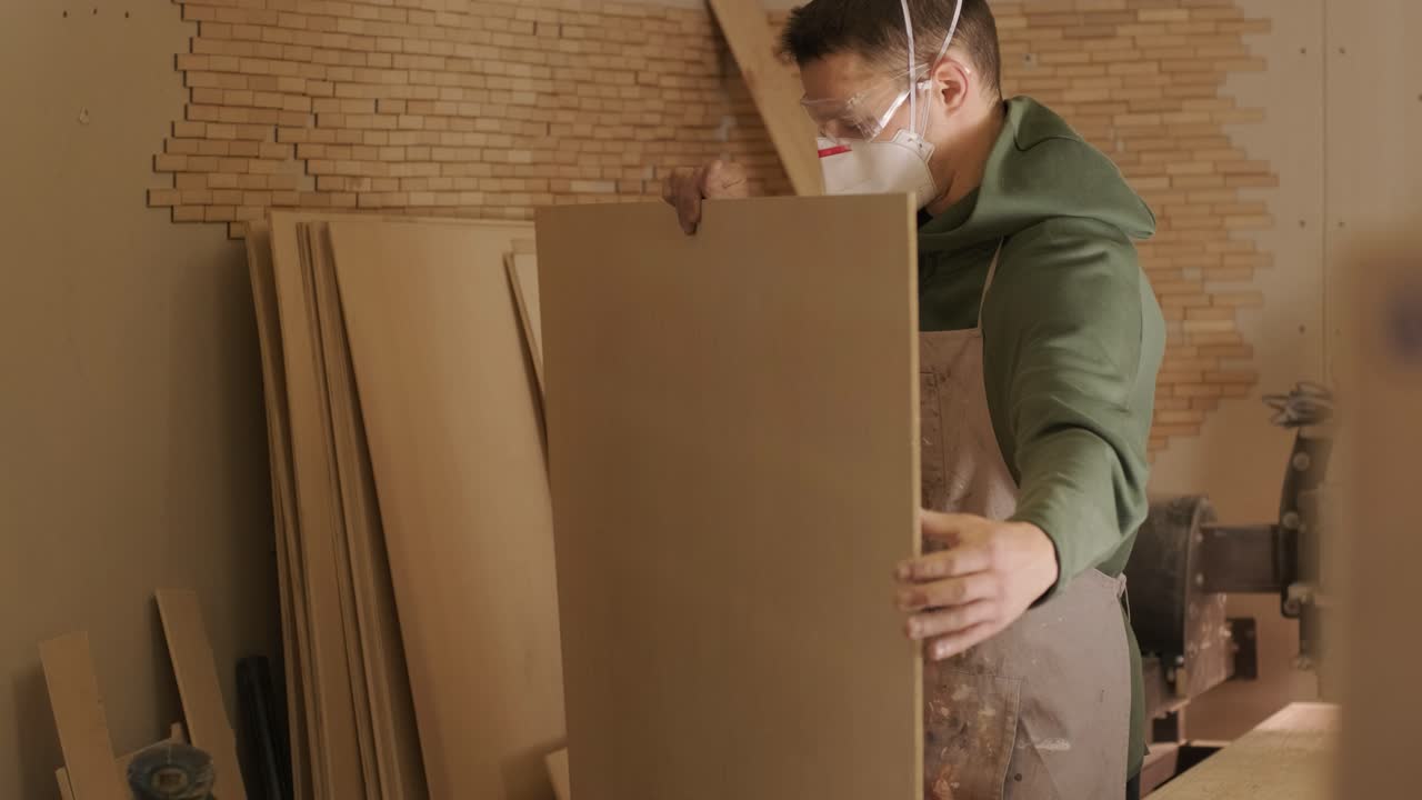 Un falegname professionista con mascherina e occhiali controlla legno grezzo, un'asse di legno e un pannello di compensato.