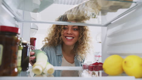 view looking out from inside of refrigerator as woman opens door and unpacks shopping bag of food