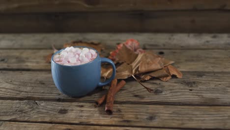 cup of hot chocolate with marshmallows, autumn leaves and cinnamon sticks on wooden surface