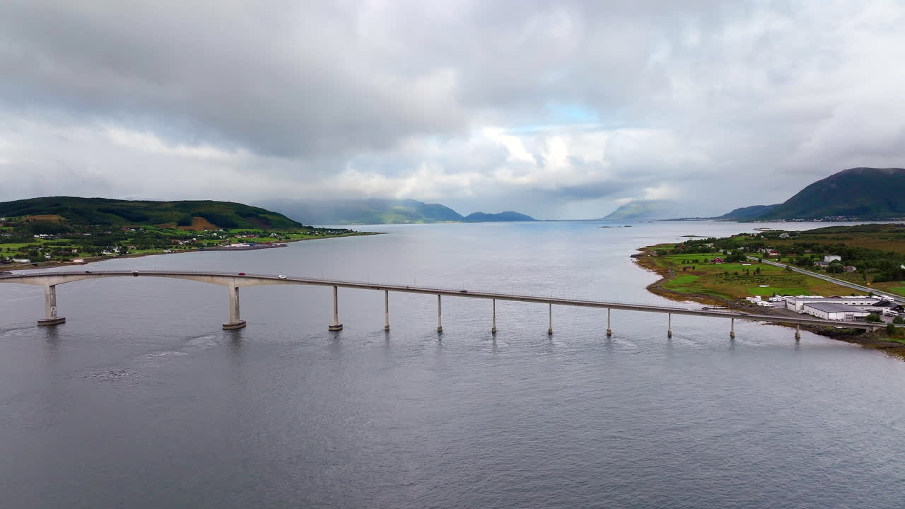 Aerial drone view of cars driving across the Sortland Bridge in Vesterålen, Norway. The concrete structure spans the fjord connecting the islands under a moody cloudy sky