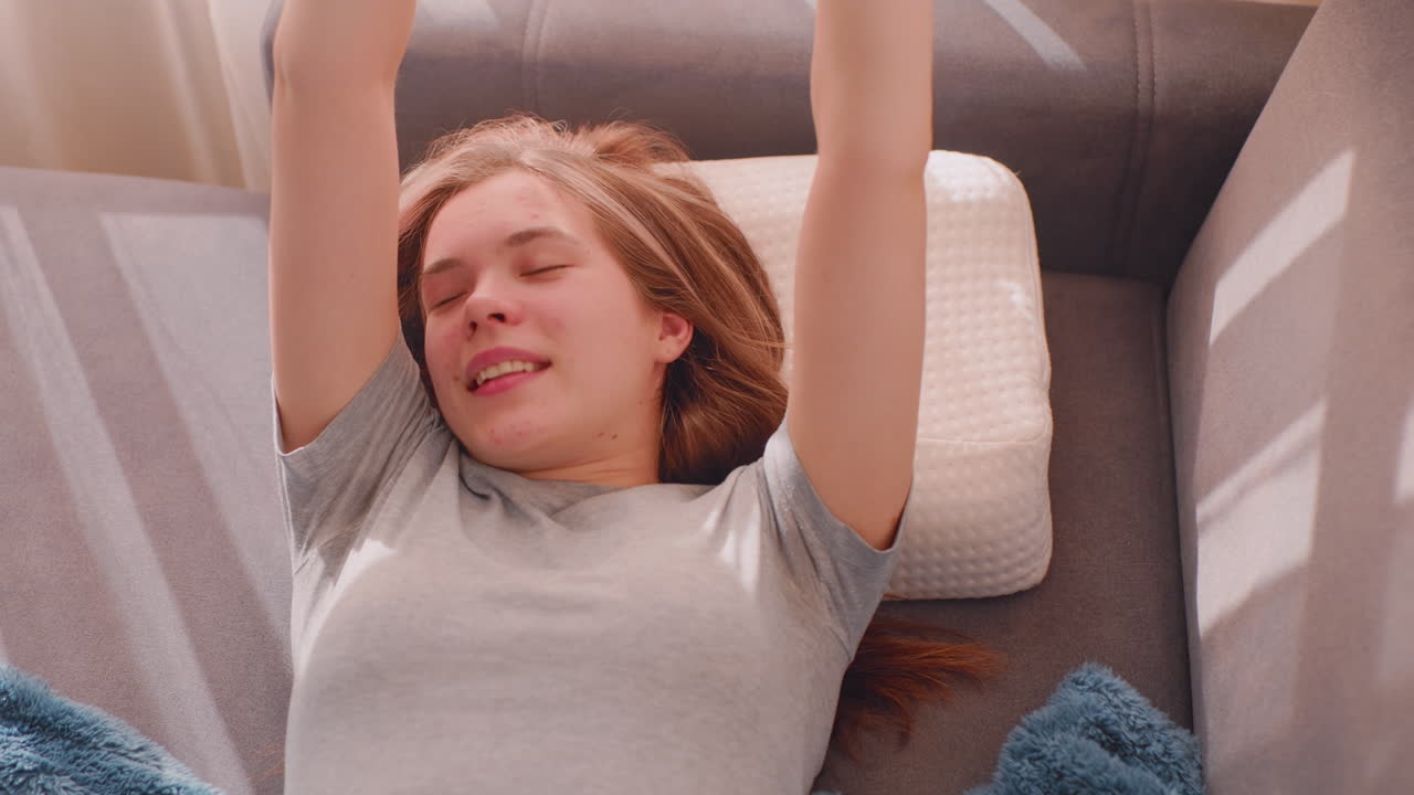 Sleepy girl finally wakes up stretching herself on couch holding soft blanket, eyes closed with relaxed expression as warm morning sunlight fills room