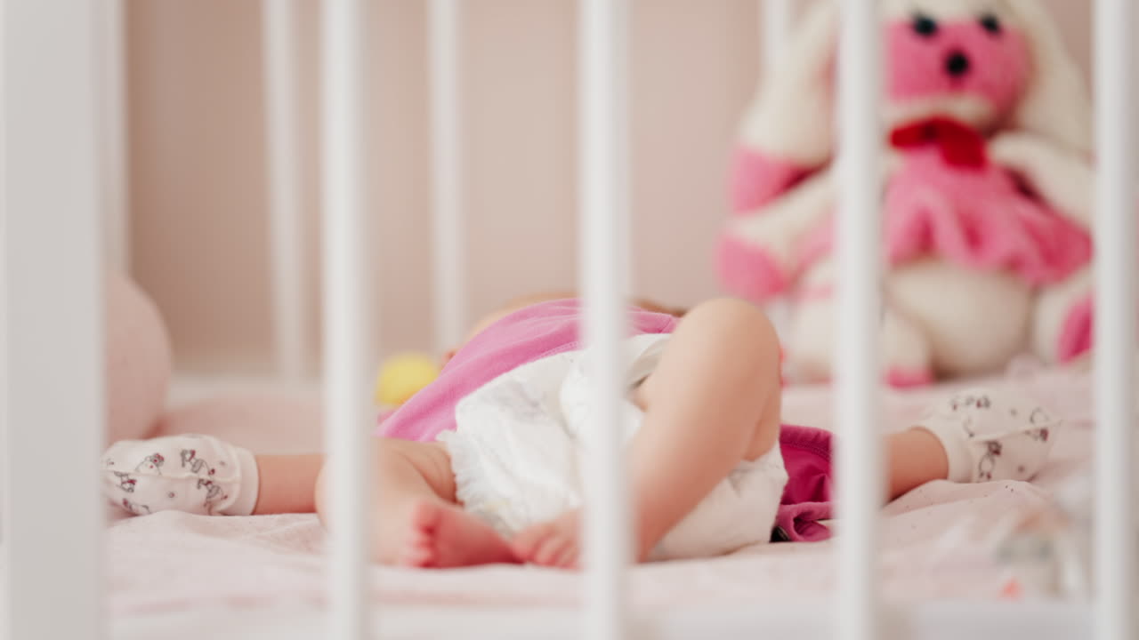 Close up of a baby's legs and diaper as the infant rests in a crib, seen through the crib bars