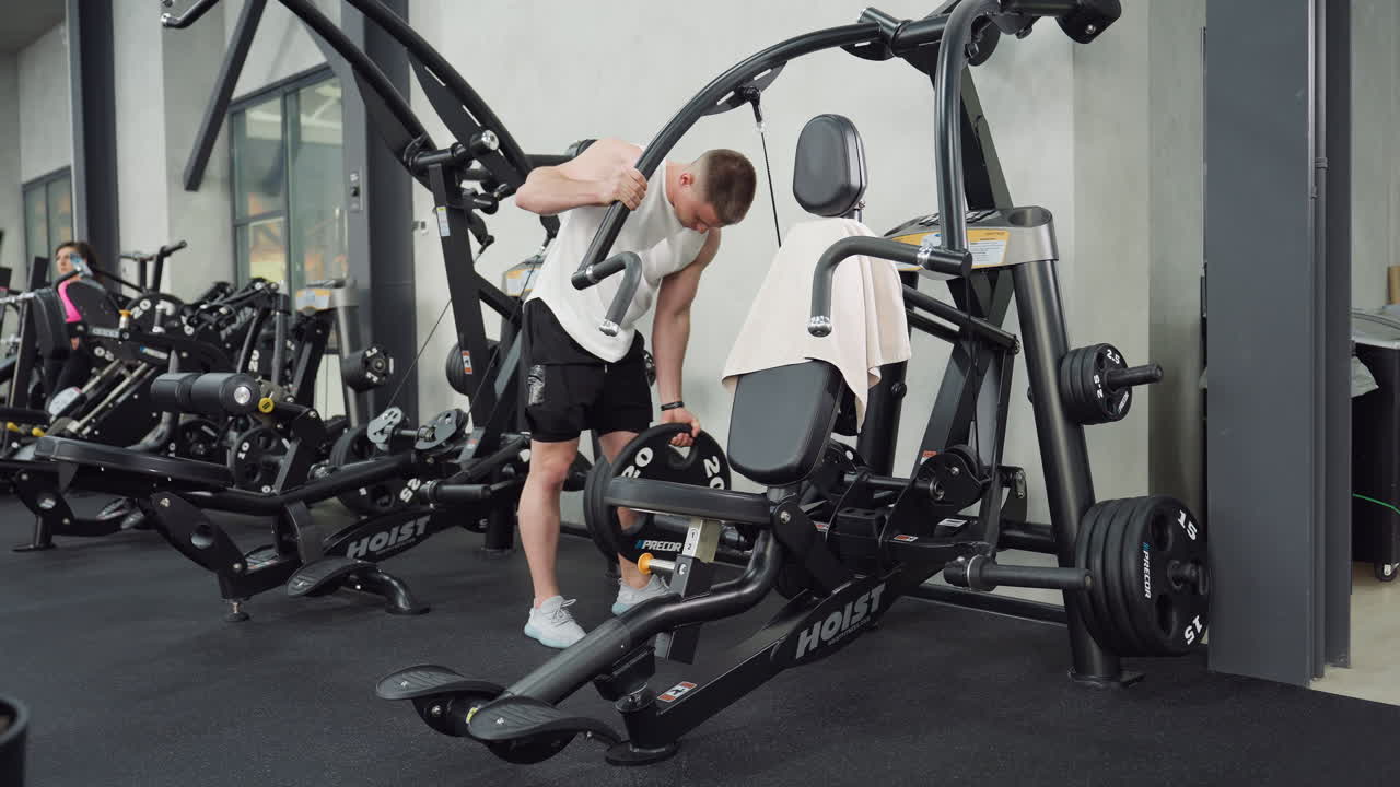 Muscular man fixes weight wheel on machine, hand turning steel plate into position during workout setup, showcasing strength and focus in modern gym environment with sleek equipment