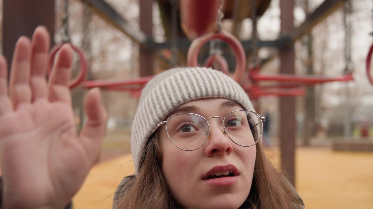 White woman inspecting hanging rings in playground, wearing beanie and glasses, winter coat and curious expression, reaching toward metal rings, quiet urban park with bare trees, muted color palette