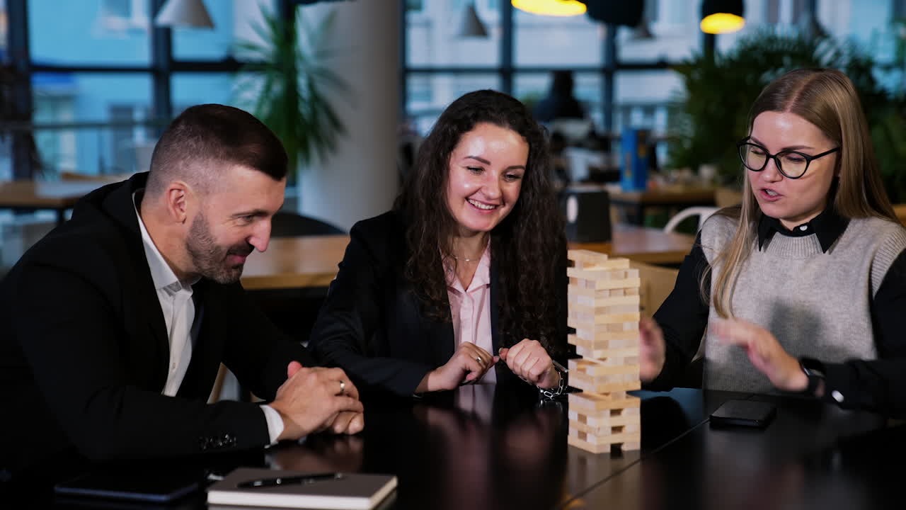 Two ladies and one man sitting at table playing table game. Male player tries to pull the brick. Blonde lady takes phone to take a picture.