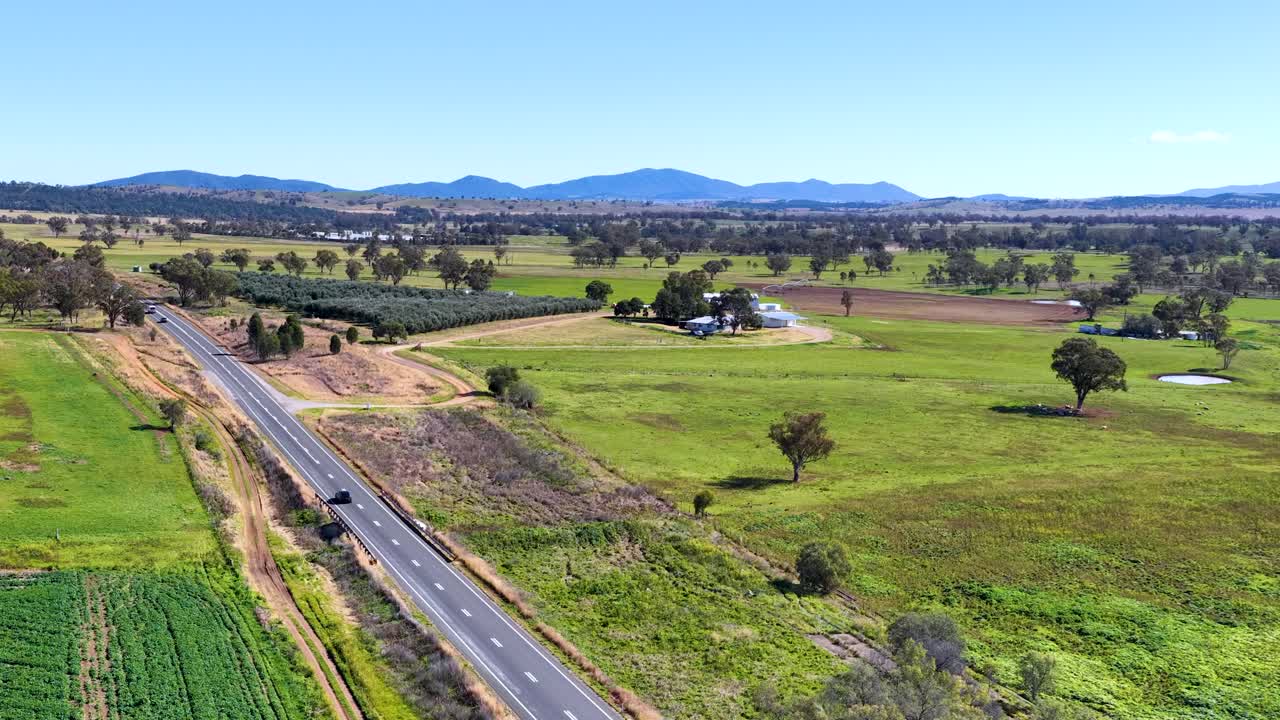 Drone camera glides above a country road and green farmland, revealing scattered trees, distant hills, and a farmhouse under bright daylight