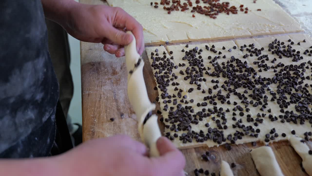 Primo piano di un fornaio che torce a mano l'impasto con gocce di cioccolato, formando fili di pasta e posizionandoli su una superficie di lavoro in legno all'interno di una panetteria tradizionale
