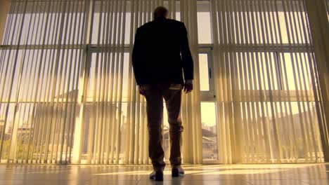 low angle dolly shot of a man walking in slow motion towards a large window where you can see a city at sunset