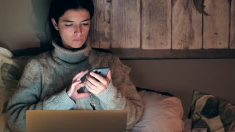 joven mujer caucásica navegando en el teléfono y la computadora portátil mientras está acostada en la cama en casa