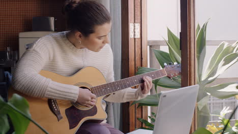 Mujer-Joven-Tocando-La-Guitarra-En-Casa-Aspirante-A-Músico-Aprendiendo-A-Tocar-Instrumentos-Musicales-Usando-Una-Computadora-Portátil-Disfrutando-De-La-Expresión-Creativa-Practicando-Música