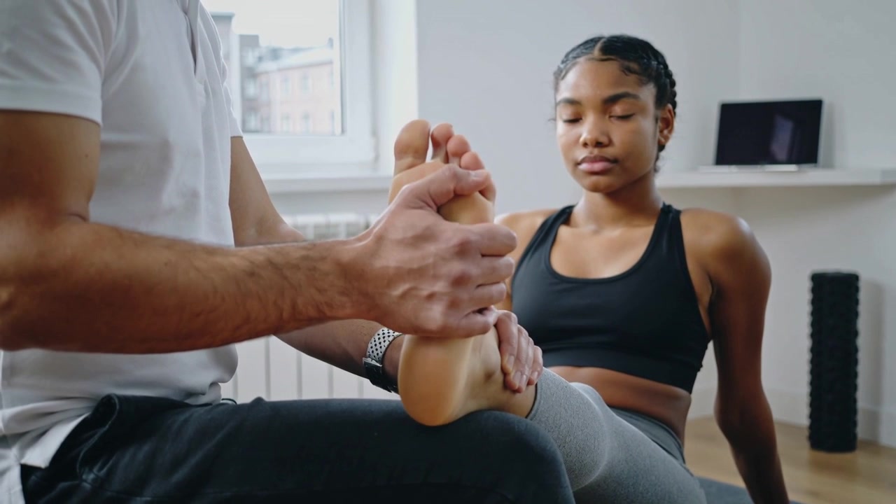 Trainer hebt, tastet und mobilisiert Fuß auf Matte zur Therapie im Heim-Fitnessstudio, Klientin in Sportkleidung.